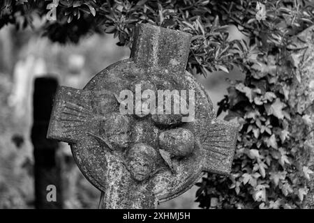 Un B&W immagine ravvicinata di una croce con edera e vegetazione alle spalle, con un cerchio di cherubini o teste di angeli al centro. Highgate Cemetery, Londra, Regno Unito Foto Stock