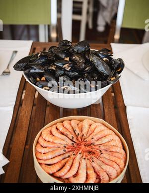 Una ciotola di cozze fresche e un piatto di gamberetti sono disposti su un tavolo da pranzo in legno all'aperto. La scena è ambientata per una festa a base di pesce, catturando il verde Foto Stock