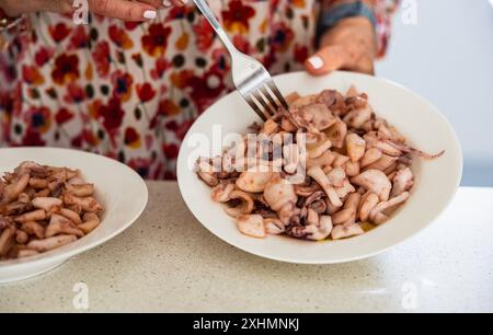 Primo piano di una donna anziana in una camicetta floreale che serve calamari cotti su un piatto, evidenziando le sue abilità culinarie e un'atmosfera familiare. Foto Stock