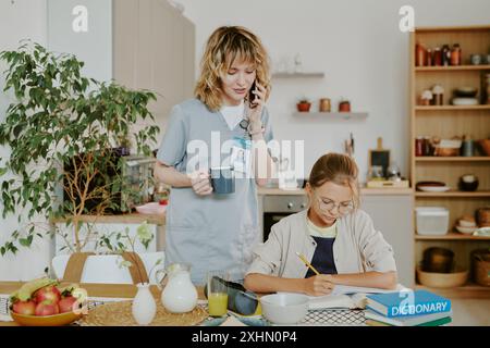 La madre del dottore controlla i compiti delle figlie e parla al telefono mentre la figlia fa esercizio nella zona pranzo Foto Stock