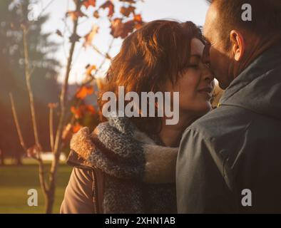 felice coppia di sposi di oltre 50 anni, camminando nel parco in autunno in una calda giornata di sole, marito che baciava la moglie sul naso, trascorrendo un giorno libero Foto Stock