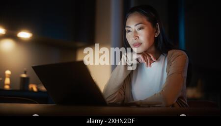 Bella e autentica donna asiatica seduta a un tavolo in un'accogliente cucina e che usa il computer portatile a casa di notte. Donna sorridente e chiacchierata sui social media, facendo shopping online, controllando le notizie Foto Stock
