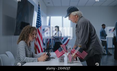 Un cittadino americano maturo del Caucaso si trova vicino al tavolo di registrazione al polling station. Una donna che ha votato parla con un uomo, usa un computer tablet e dà le schede elettorali per votare. Il giorno delle elezioni negli Stati Uniti. Foto Stock