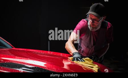 Esperto di autolavaggio che utilizza un panno in microfibra per asciugare una moderna Sportscar rossa. Uomo adulto pulizia della sporcizia, preparazione di una muscle car americana per il dettaglio. Foto cinematografica creativa con veicolo sportivo Foto Stock