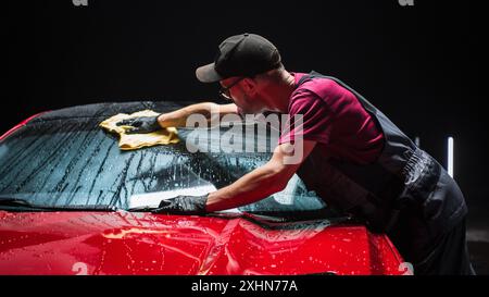 Esperto di autolavaggio che utilizza un panno in microfibra per asciugare una moderna Sportscar rossa. Uomo adulto pulizia della sporcizia, preparazione di una muscle car americana per il dettaglio. Foto cinematografica creativa con veicolo sportivo Foto Stock