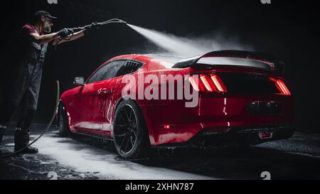 Esperto di autolavaggio che utilizza l'idropulitrice ad acqua per pulire una moderna Sportscar rossa. Uomo adulto che lava via la sporcizia, prepara un'auto muscolare americana per il dettaglio. Riprese cinematografiche creative con veicolo di lusso Foto Stock