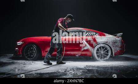 Esperto di autolavaggio che utilizza l'idropulitrice ad acqua per pulire una moderna Sportscar rossa. Uomo adulto lavando via lo shampoo, preparando una Muscle Car per i dettagli. Foto creative dal basso con veicolo sportivo Foto Stock