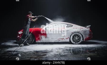 Esperto di autolavaggio che utilizza l'idropulitrice ad acqua per pulire una moderna Sportscar rossa. Uomo adulto che lava via la sporcizia, prepara un'auto muscolare americana per il dettaglio. Foto cinematografiche creative con veicolo di lusso Foto Stock