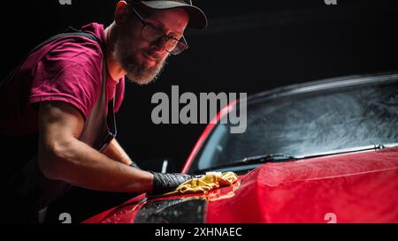 Esperto di autolavaggio che utilizza un panno in microfibra per asciugare una moderna Sportscar rossa. Uomo adulto pulizia della sporcizia, preparazione di una muscle car americana per il dettaglio. Foto cinematografica creativa con veicolo sportivo Foto Stock