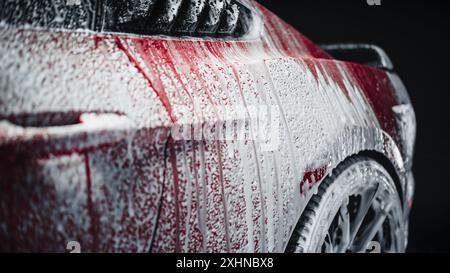 Foto estetica di una coupé sportiva rossa ricoperta di sapone e schiuma. Primo piano di schiuma che gocciola dal parafango posteriore della vettura sul volante con pneumatici e cerchioni Performance Foto Stock