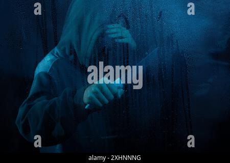 Ritratto di un uomo misterioso che indossa una felpa dietro un vetro bagnato e tiene in mano un coltello. Faccia nascosta. Ritratto in studio. Isolato su sfondo blu. Foto Stock