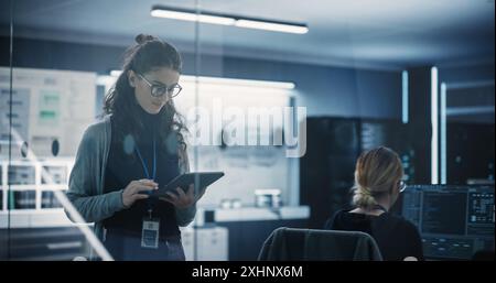 Responsabile del dipartimento di ingegneria del software tecnologico camminare e utilizzare un tablet. Donna multietnica che lavora in ufficio Foto Stock