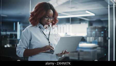 Ritratto di un responsabile del dipartimento di ingegneria del software tecnologico che sta in piedi e utilizza un computer portatile. Donna africana che guarda in lontananza e pensa mentre lavora in ufficio Foto Stock