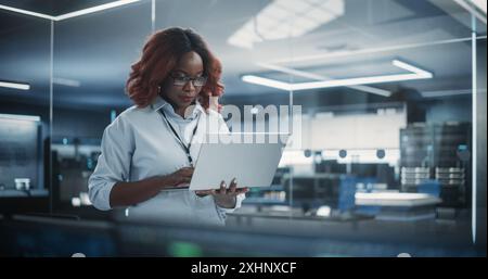 Ritratto di un responsabile del dipartimento di ingegneria del software tecnologico che sta in piedi e utilizza un computer portatile. Donna africana che guarda in lontananza e pensa mentre lavora in ufficio Foto Stock