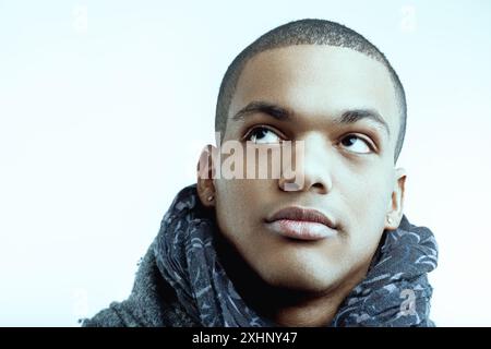 Uomo con cappotto grigio e sciarpa a motivi blu che tocca il mento con attenzione. I suoi capelli corti sono ben rifiniti, e guarda verso l'alto, riflettendo un momen Foto Stock