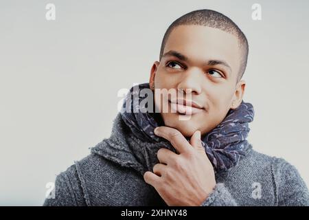 Giovane con un'espressione premurosa, toccando il mento con la mano destra. È vestito con un cappotto di lana grigia e una sciarpa a motivi blu, che ti guarda Foto Stock