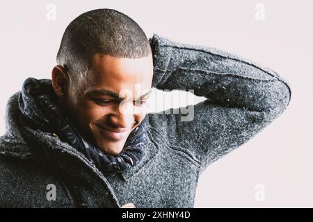 Giovane con un sorriso timido, guardando in basso e toccando la parte posteriore della testa con la mano sinistra. Vestito con un cappotto di lana grigia e sciarpa blu, il suo espresso Foto Stock