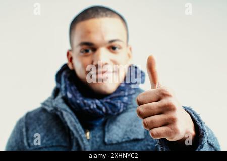 Giovane uomo (sfocato) con un'espressione amichevole, che dà un gesto di messa a fuoco con il pollice. È vestito con un cappotto di lana grigio e una sciarpa blu, concentrandosi Foto Stock