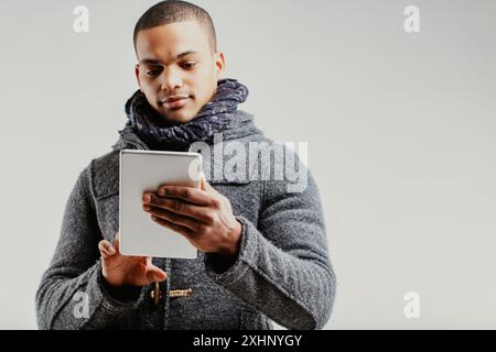 Uomo con un cappotto grigio e sciarpa blu, che utilizza un tablet digitale per essere sempre informato. La sua seria espressione mette in evidenza l'impegno per la conoscenza e la comprensione Foto Stock