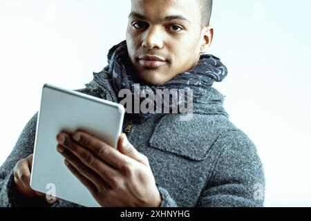 Giovane con un'espressione seria, con un tablet digitale e sempre informato. Vestito con un cappotto di lana grigio e sciarpa blu, si concentra sulla conoscenza Foto Stock