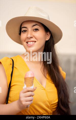 Giovane donna con un cappello e un vestito giallo sorridente mentre tiene in mano un cono gelato. Le piace una delizia rinfrescante e appare allegra in una giornata di sole. Foto Stock