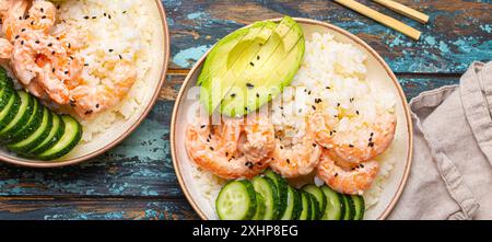 Due ciotole di ceramica bianca con riso, gamberetti, avocado, verdure, semi di sesamo e bacchette su un colorato sfondo rustico in legno con vista dall'alto Foto Stock