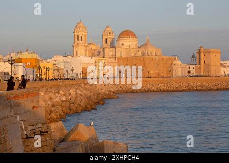 Spagna, Andalusia, Cadice, paseo maritimo, passeggiata costiera accogliendo passeggini lungo le facciate di vecchi edifici e la cattedrale in luce calda Foto Stock