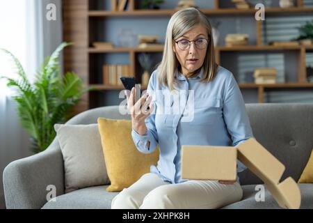 Donna anziana che prova sorpresa e confusione mentre sboccia un pacchetto inaspettato e usa lo smartphone, seduto sul divano in salotto. Pianta verde e libreria sullo sfondo. Foto Stock