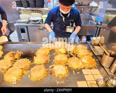 Chef che preparano okonomiyaki, conosciuto come il cibo dell'anima di Hiroshima e servito in circa 2.000 ristoranti in e intorno alla città. L'okonomiyaki apparve per la prima volta dopo la seconda guerra mondiale come cibo a buon mercato e riempitivo per soddisfare la fame dei lavoratori. Il piatto comprende carne di maiale, uova e spaghetti soba. Realizzata con uno strato di base sottile e crepe e amalgamata di ingredienti, la frittella viene cotta su una piastra lunga e piatta. La polpa contiene scaglie di alghe verdi essiccate, cipolle verdi, germogli di soia e cavolo. Giappone. Foto Stock