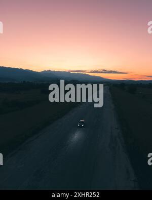 Panorama verticale di un'auto con luci accese in una grande strada vuota con un bellissimo tramonto sullo sfondo. Foto Stock
