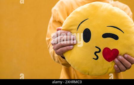 Una persona che regge un cuscino giallo di emoji con una faccia di bacio su uno sfondo giallo, con uno spazio per le copie Foto Stock