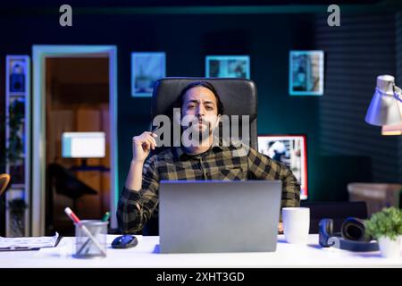 Ritratto di un uomo barbuto comodamente seduto alla sua scrivania, tenendo una penna e guardando la fotocamera. Il freelance maschile sembra serio e si sta preparando ad usare il suo portatile per lavoro. Foto Stock