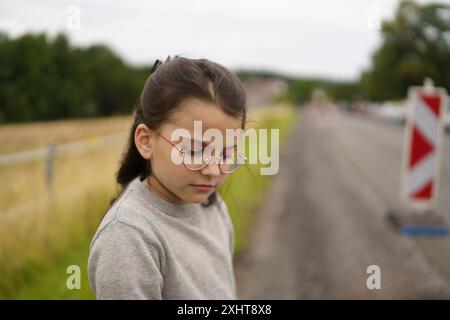 Una studentessa bambina che indossa gli occhiali sullo sfondo di una strada asfaltata e di erba verde. Concetto di viaggio per bambini Foto Stock