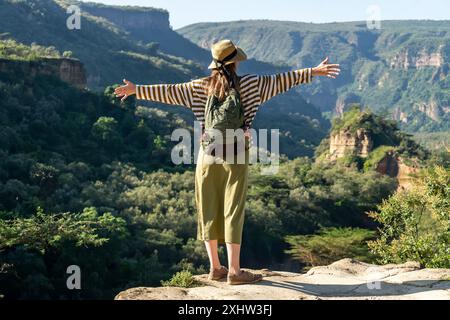 Vista posteriore di una donna entusiasta in un cappello con uno zaino con le mani alzate, guardando dall'alto un paesaggio di montagna incredibilmente bello Foto Stock