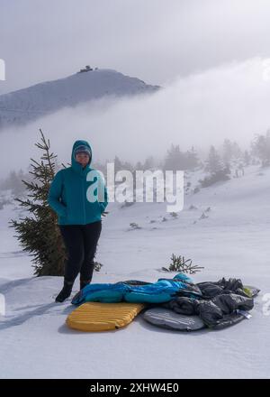 Una donna di mezza età si erge sullo sfondo di montagne innevate. Di fronte a lei, i sacchi a pelo e i materassi giacciono sulla neve. Viaggi invernali Foto Stock