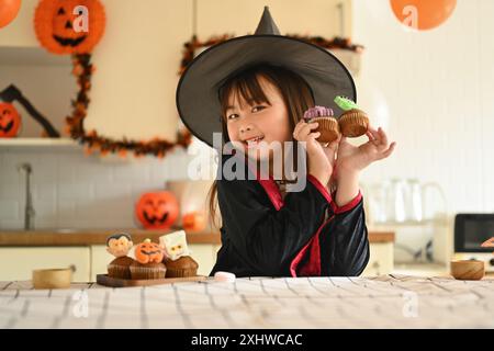 Ritratto della dolce bambina in costumi da strega che reggono i cupcake di Halloween in piedi in una stanza decorata Foto Stock