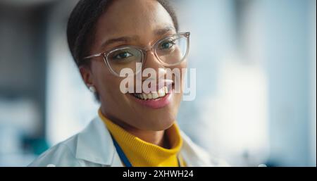 Ritratto di giovane donna nera sorridente che indossa occhiali e camice da laboratorio che guarda la fotocamera. Futuro ingegnere che persegue la carriera scientifica. Industrial Manufacturing Student in University Laboratory Posing Foto Stock