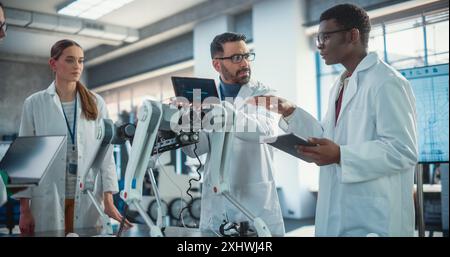 Team diversificato di specialisti di robotica industriale che lavorano su un robot mobile. Giovani ingegneri in camici da laboratorio che discutono di un cane robotico automatizzato ai con il loro supervisore di progetto, utilizzando un tablet digitale Foto Stock