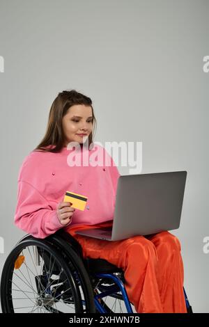 Una giovane donna in sedia a rotelle siede in un monolocale, utilizzando un computer portatile e tenendo in mano una carta di credito. Foto Stock