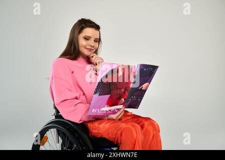 Una giovane donna bruna in un abbigliamento casual si siede su una sedia a rotelle e legge una rivista in un ambiente studio. Foto Stock