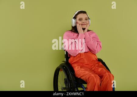 Una giovane donna bruna in sedia a rotelle ascolta la musica con le cuffie in un ambiente da studio. Indossa un maglione rosa e pantaloni arancioni. Foto Stock