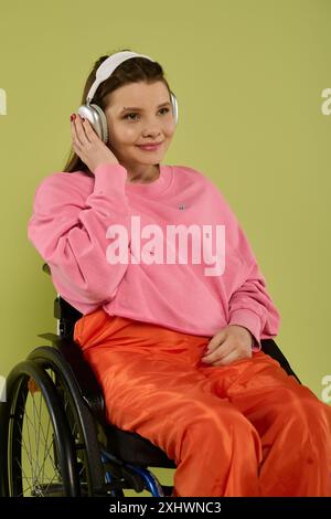 Una giovane donna bruna siede su una sedia a rotelle in un ambiente da studio, indossando un maglione rosa e pantaloni arancioni. Sta ascoltando musica con le cuffie, e. Foto Stock