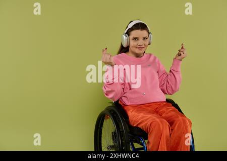 Una giovane donna con i capelli marroni siede su una sedia a rotelle in un ambiente da studio, indossando un maglione rosa e pantaloni arancioni. Indossa le cuffie e sorride come se fosse Foto Stock