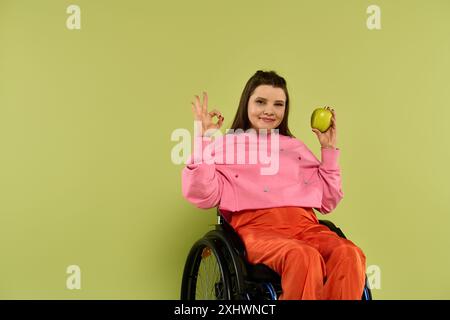 Una giovane donna bruna in sedia a rotelle siede in un ambiente da studio, indossando un abbigliamento casual. Tiene una mela verde in una mano e fa un segno di OK con Foto Stock