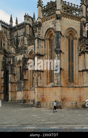 Il bambino guida uno scooter autobilanciato passando davanti alla facciata gotica del monastero di batalha, mettendo in evidenza i suoi torreggianti archi e i dettagli intricati Foto Stock