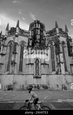 Un giovane ciclista con un casco rosso passa davanti alla facciata gotica del monastero di Batalha, mostrando le sue intricate opere in pietra Foto Stock