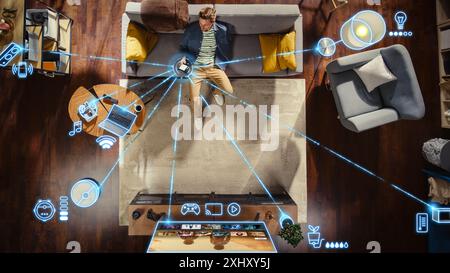 Vista dall'alto dell'uomo caucasico nell'appartamento mansardato seduto sul divano e collegando lo smartphone al comodo sistema Smart Home. Modifica VFX per visualizzare i dispositivi connessi. Laptop, TV, altoparlante. Foto Stock