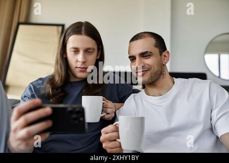 Una giovane coppia gay si gode una mattinata accogliente insieme nella loro camera da letto, condividendo un drink caldo e guardando il telefono. Foto Stock