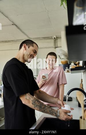 Due giovani uomini, in una casa moderna, si godono un momento mattutino informale mentre uno lava i piatti mentre l'altro tiene una tazza. Foto Stock