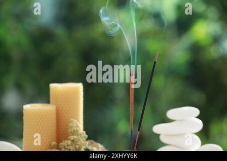 Bastoncini di incenso che si muovono vicino a pietre, candele e fiori secchi su sfondo verde sfocato Foto Stock